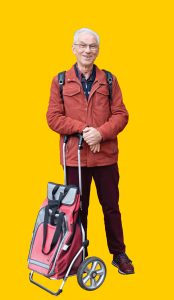04 Gelber Hintergrund, davor ein Mensch im Portrait im Rucksack und Ziehwagen