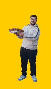 06 Gelber Hintergrund, davor ein Mensch im Portrait, in der Hand ein Blech voller Baklava